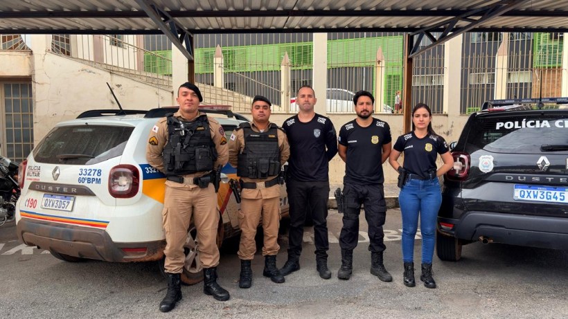 Cinco policiais em uniformes, posando em frente a dois veículos policiais estacionados.