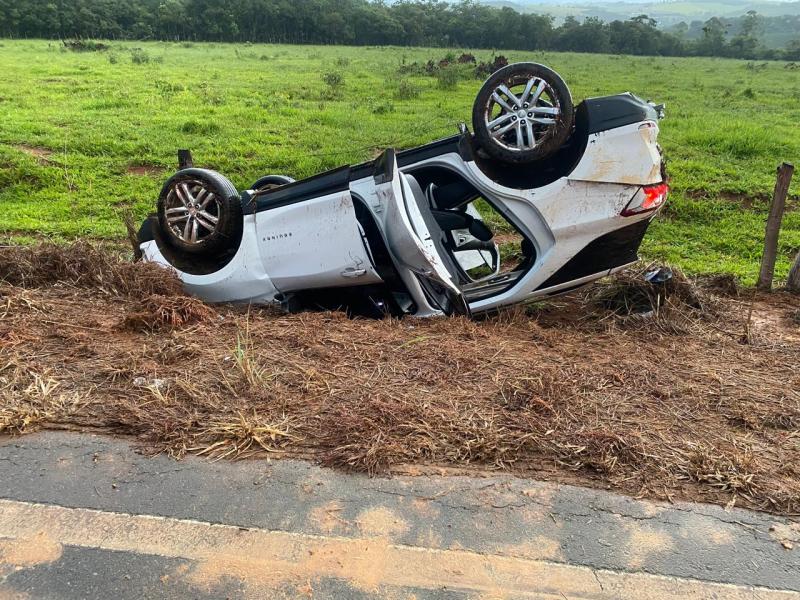 Carro branco capotado ao lado da estrada, com pneus para cima, em um ambiente rural com vegetação ao fundo.