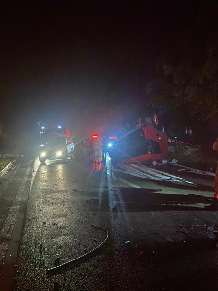 Acidente de carro à noite, com um veículo capotado na pista e viaturas de emergência na cena.