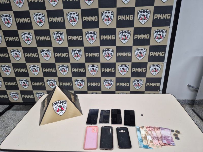 Mesa com vários celulares, dinheiro em notas e moedas, e uma pirâmide com o logotipo do PMMG ao fundo.