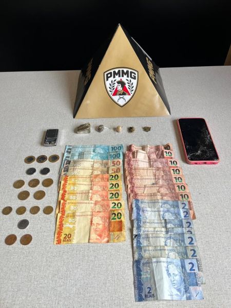 Imagem com uma pirâmide dourada com o logotipo PMMG, acompanhada por notas e moedas de reais, um celular rosa com tela danificada e pequenas pedras em uma mesa.