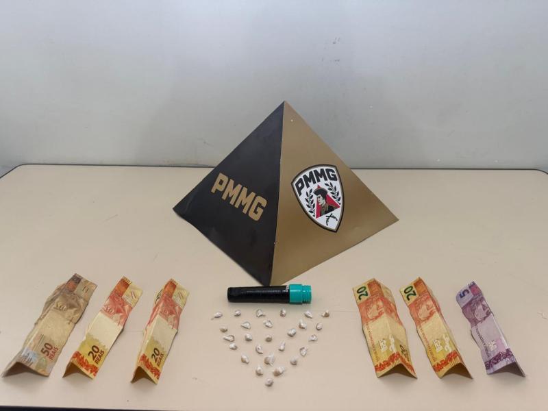 Uma pirâmide com as inscrições PMMG, acompanhada de notas de dinheiro (20, 50 e 5 reais) e um pequeno objeto preto com tampa azul, além de pequenas pedras brancas dispostas em forma de triângulo.