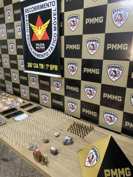Mesa com diversos itens apreendidos, incluindo pedras e objetos não identificáveis, em frente a um fundo com logotipos da Polícia Militar, sinalizando uma operação policial.
