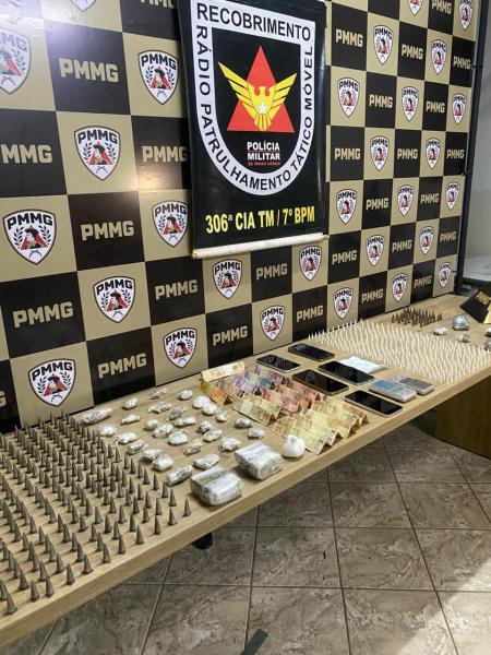 Mesa com uma variedade de drogas e munições expostas, com fundo de uma parede decorada com logotipos da Polícia Militar de Minas Gerais.