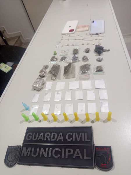 Mesa com diversos itens apreendidos, incluindo pacotes de substâncias, equipamentos eletrônicos e insígnias da Guarda Civil Municipal.