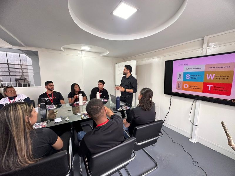 Reunião em sala com pessoas discutindo análise SWOT, apresentada por um homem em pé diante de um grupo.