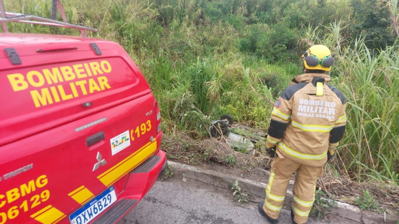 Um bombeiro militar, vestindo um traje de segurança, observa uma área verde ao lado de um veículo de emergência identificado como "BOMBEIRO MILITAR".