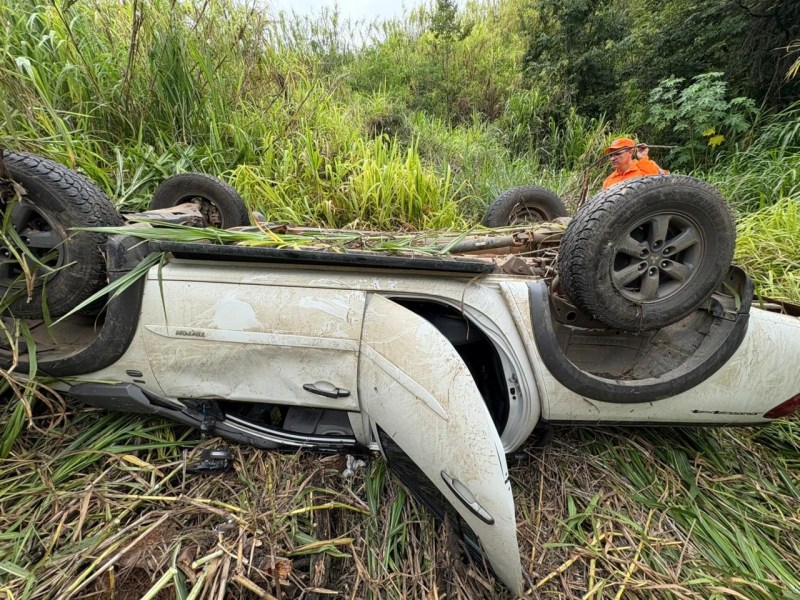 Carro capotado em vegetação alta, com rodas para cima e lama visível, enquanto um homem em roupas laranjas observa ao fundo.