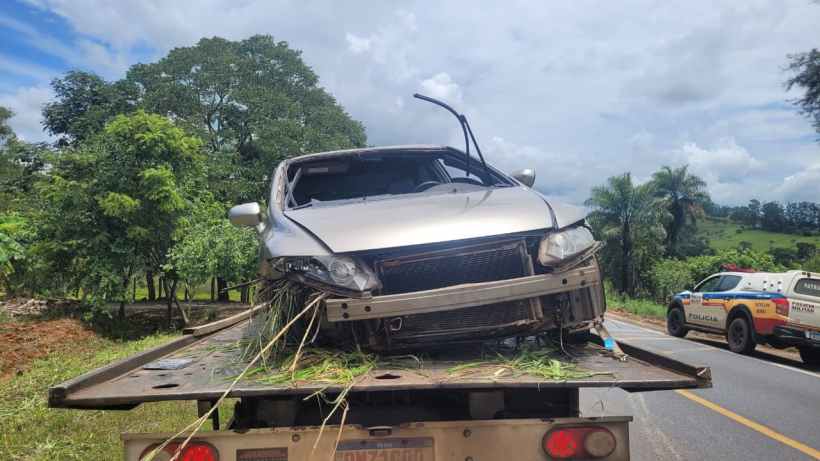 Um carro danificado sendo rebocado em uma estrada, com árvores e vegetação ao fundo.