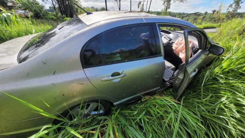 Carro acidentado em área de grama alta, com a porta aberta e visíveis sinais de danos na lateral.