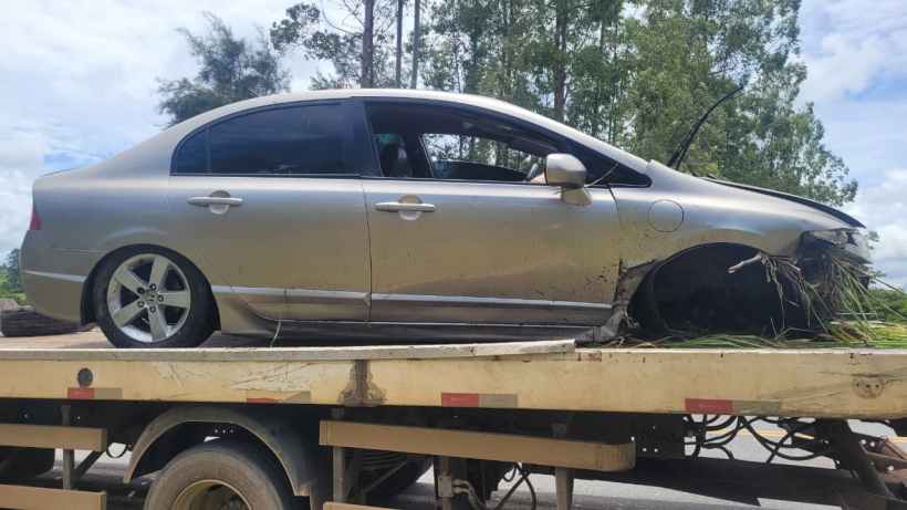 Um carro prata danificado sendo transportado em uma plataforma, com vegetação ao redor.
