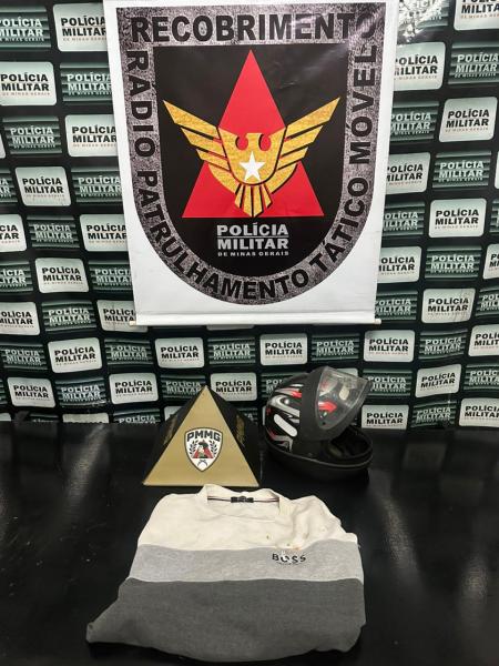 Imagem com uma camiseta da marca Boss, um capacete e um objeto em formato de pirâmide, sobre uma superfície preta, com um fundo de banner da Polícia Militar.