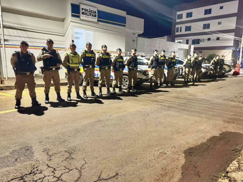 Grupo de policiais militares em frente à sede da polícia à noite, posando para a foto ao lado de viaturas.
