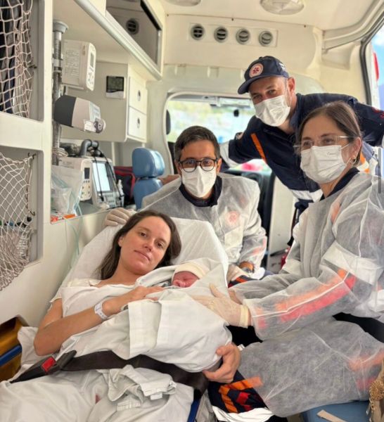 Mulher segurando um bebê recém-nascido em uma ambulância, cercada por profissionais de saúde, todos usando máscaras e equipamentos de proteção.