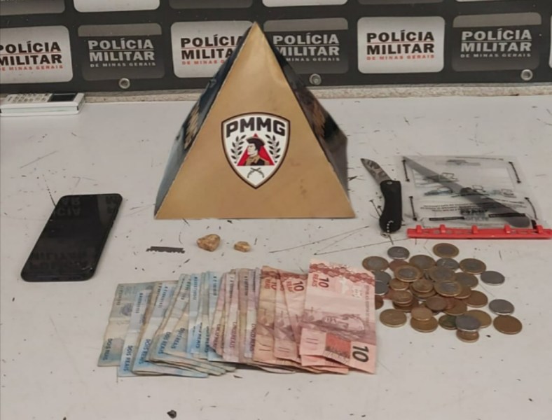 Imagem de uma mesa com itens apreendidos pela polícia, incluindo uma pirâmide com o logo da Polícia Militar de Minas Gerais, um celular preto, uma faca, notas de dinheiro e moedas.