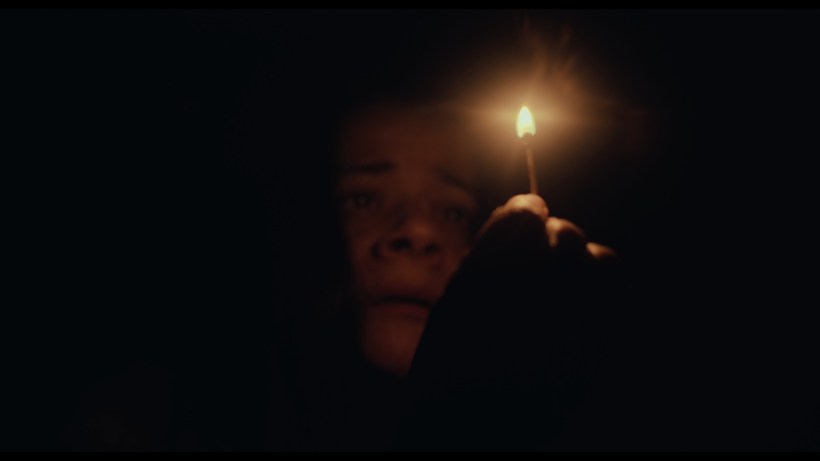 Pessoa segurando uma vela acesa em um ambiente escuro, com uma expressão de medo ou preocupação.