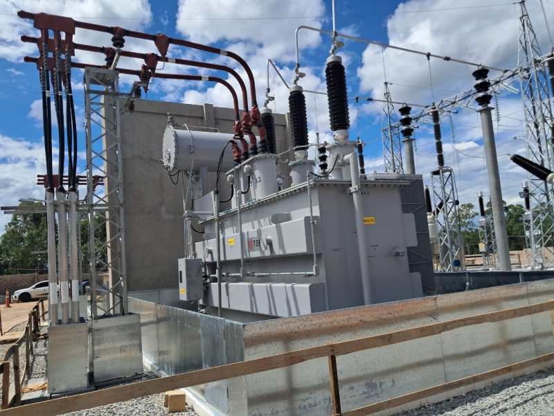 Transformador em estação elétrica com estruturas de transmissão e céu parcialmente nublado.