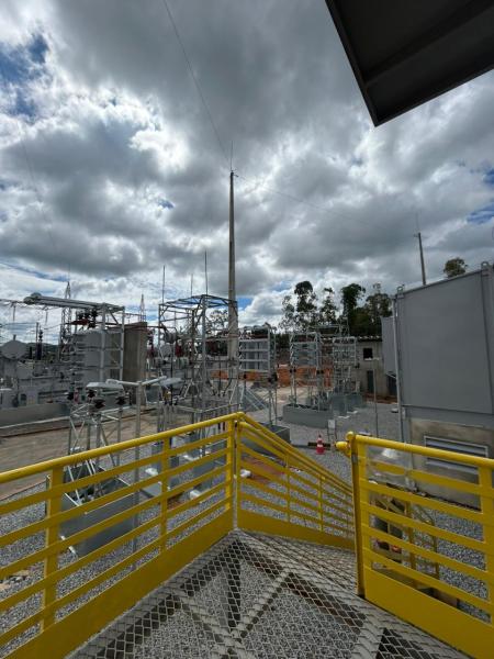 Vista de uma subestação elétrica com equipamentos altos e estrutura metálica, sob um céu nublado, com uma escada amarela em primeiro plano.