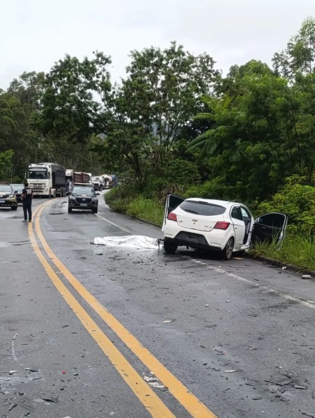 Acidente de carro em uma estrada, com um veículo branco parado na lateral e destroços espalhados na pista. Caminhões e carros estão parados nas proximidades.
