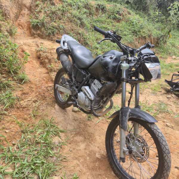 Motocicleta preta estacionada em um terreno de barro, cercada por vegetação.