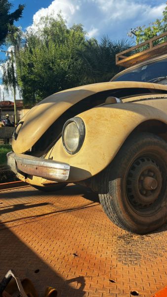 Um carro Fusca amarelo antigo com capô levemente levantado, estacionado em uma superfície de metal laranja. O fundo apresenta árvores e céu parcialmente nublado.