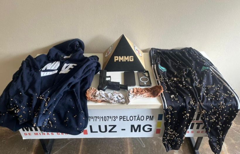 Conjunto de roupas incluindo um moletom e calças esportivas com grãos, uma arma de fogo, um celular e outros objetos sobre uma mesa com a inscrição PMMG.