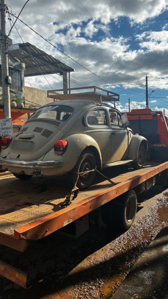 Um carro antigo, estilo fusca, estacionado em uma carroceria de caminhão, com céu nublado ao fundo.