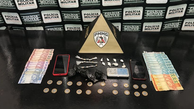 Mesa com dinheiro, celulares e drogas, incluindo embalagens e substâncias, exibidos sob um fundo com placas da Polícia Militar.