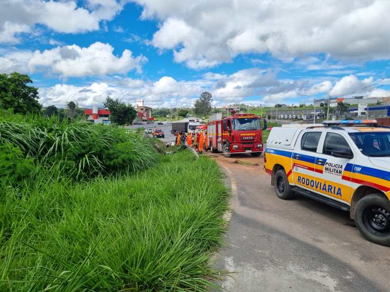 Caminho com vegetação alta e veículos de emergência, incluindo um caminhão de bombeiros e uma viatura da polícia rodoviária, em uma estrada com céu azul e nuvens.