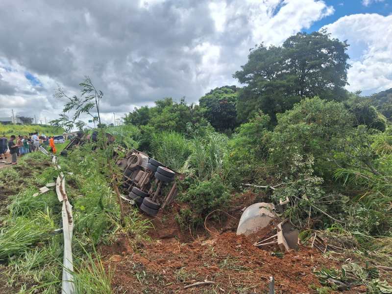 Acidente de caminhão tombado ao lado da estrada, com vegetação ao redor e céu nublado.