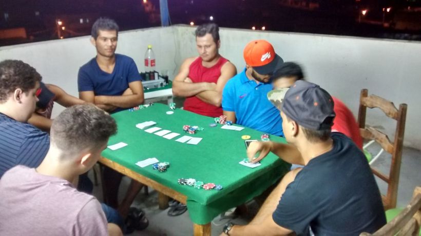 A group of men playing poker around a green felt table, with poker chips and playing cards displayed. The setting is on a balcony at night, with city lights visible in the background.