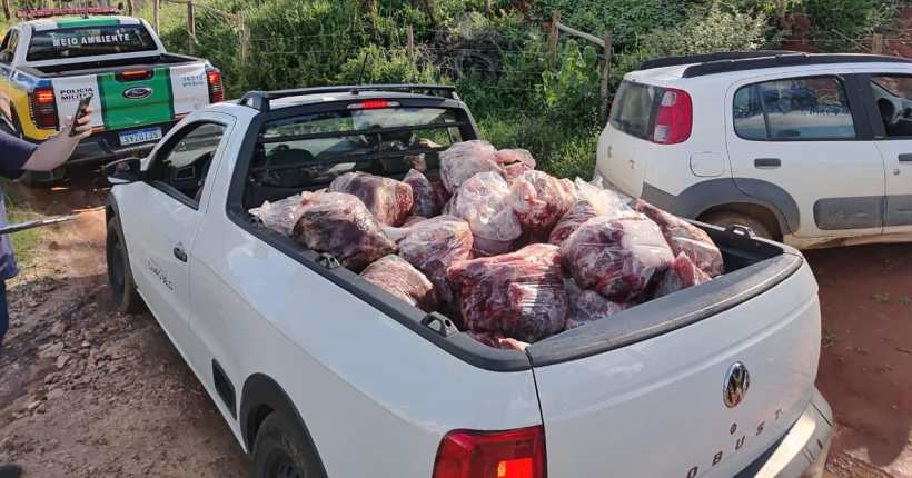 Caminhonete branca com carga de carne embalada na caçamba, estacionada em uma estrada rural, com veículos de polícia ambiental ao fundo.