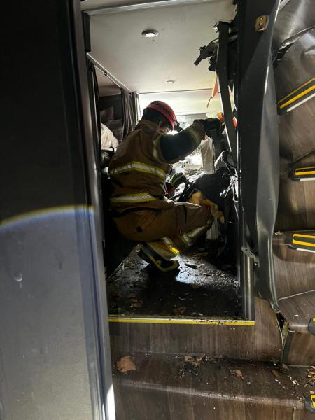 Um bombeiro em uniforme de combate a incêndio, com capacete, trabalhando dentro de um veículo danificado, cercado por destroços e com iluminação escassa.