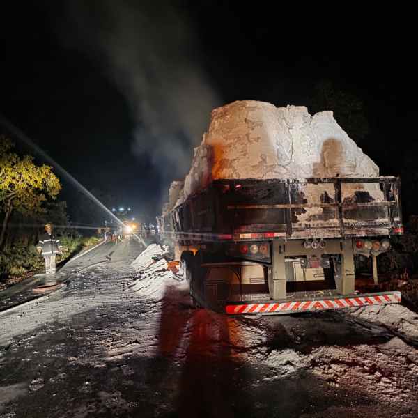 Caminhão carregado de sal tombado na estrada à noite, com fumaça visível e equipes de emergência ao fundo.