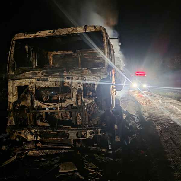 Caminhão queimado à noite, com fumaça saindo e luzes de emergência ao fundo.