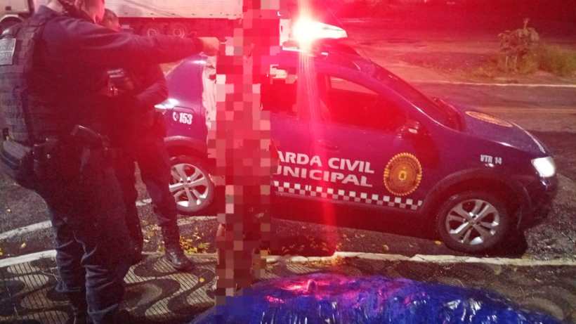 Policiais da Guarda Civil Municipal em ação noturna, com um veículo oficial ao fundo e luzes vermelhas intermitentes.