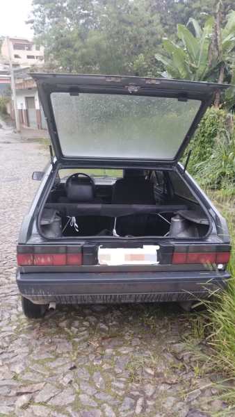 Um carro preto visto de trás, com a porta traseira aberta, exibindo o interior vazio e alguns fios visíveis. O cenário ao fundo inclui vegetação e uma estrada de pedra.