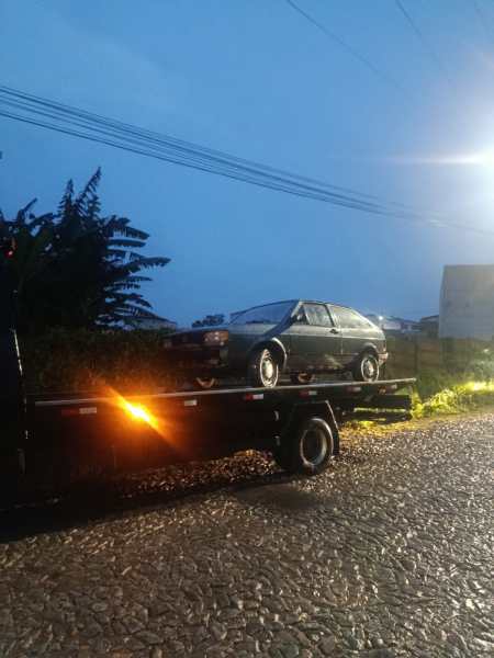 Um carro sendo transportado em um guincho à noite, com iluminação suave e um fundo de vegetação ao redor.