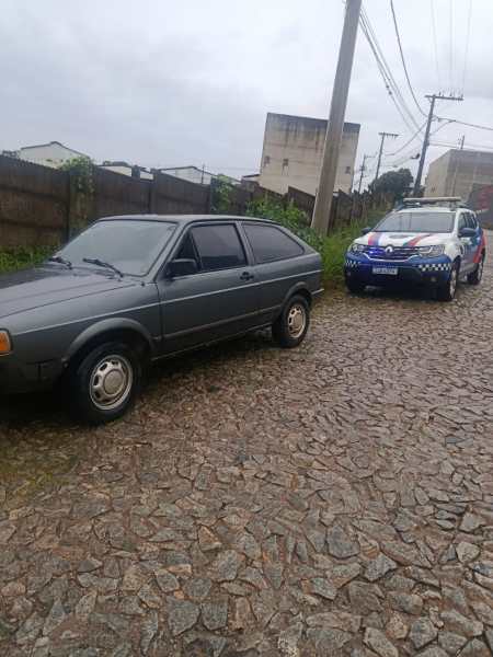 Um carro cinza estacionado em uma rua de pedras, com um veículo da polícia ao fundo.