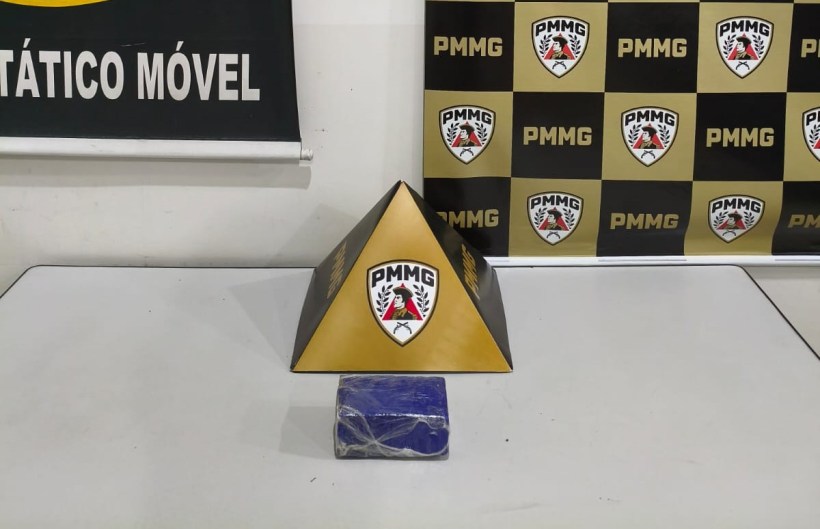 Uma pirâmide dourada com o logotipo PMMG em cima de uma mesa, ao lado de um bloco azul envolto em plástico. Ao fundo, um painel com as cores preto e dourado, também apresentando o logotipo PMMG.
