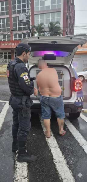Um policial abordando um homem nu na rua, próximo a um veículo policial com a porta aberta.