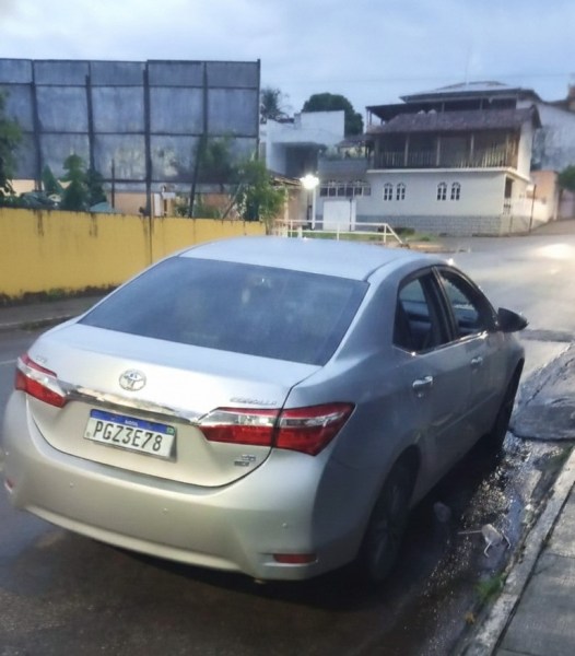 Uma imagem de um carro Toyota prata estacionado em uma rua à noite, com iluminação suave e um fundo de construções e um muro amarelo.