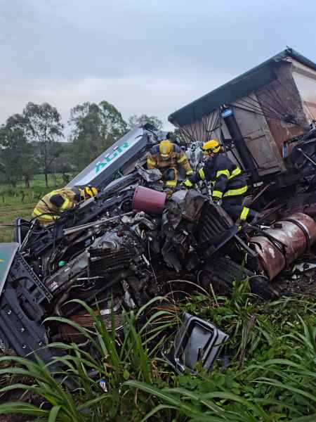 Bombeiros trabalhando em um caminhão tombado em uma área rural, com vegetação ao redor.