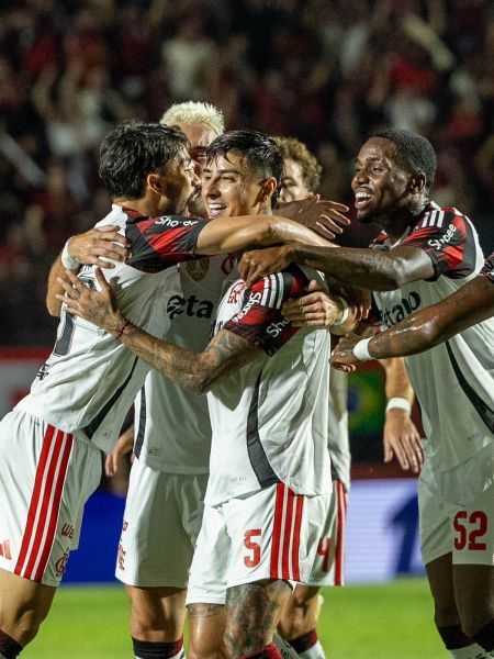 A group of soccer players celebrating a goal, embracing each other in excitement, wearing matching jerseys with red and black accents.