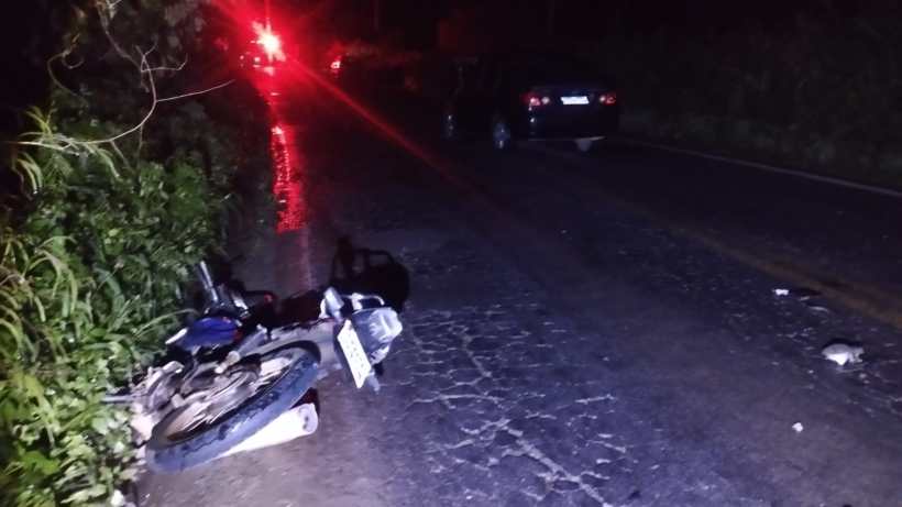 Moto caída ao lado da estrada em uma noite escura, com um carro visível ao fundo e luzes vermelhas refletindo.