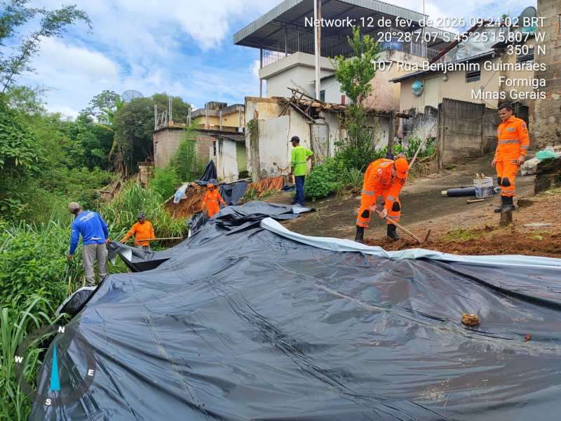 Grupo de trabalhadores em uma encosta, cobrindo o solo com lona preta, cercados por vegetação e residências em ruínas. Eles usam uniformes laranja e estão realizando atividades de contenção ou proteção no local.