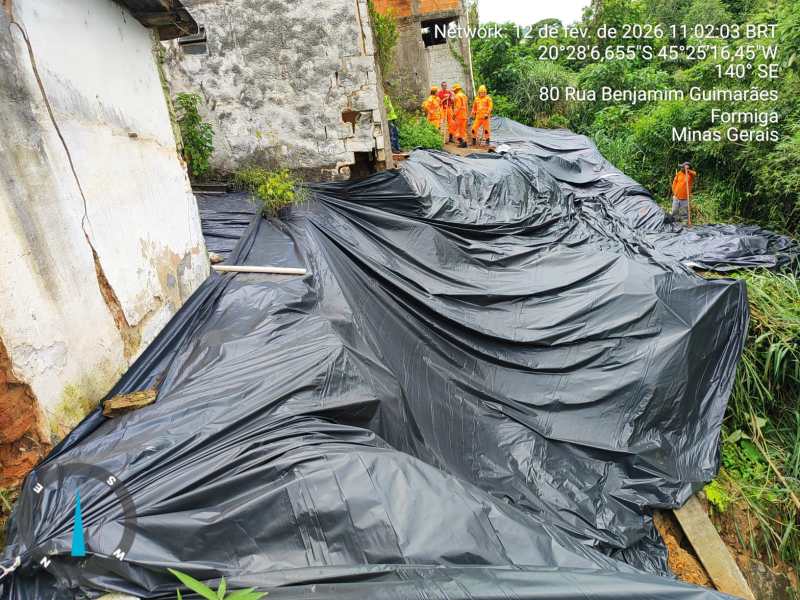 Visão de uma área coberta com lona preta, com operários em uniforme laranja trabalhando ao fundo. A cena é localizada em Formiga, Minas Gerais.