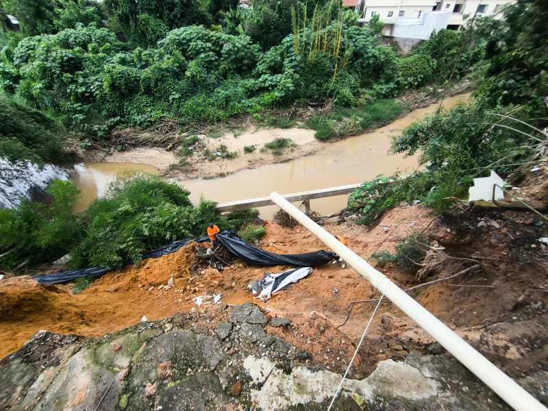 Imagem mostrando uma erosão em um terreno com encosta de terra, um rio ao fundo e um trabalhador vestido de laranja perto da área afetada.