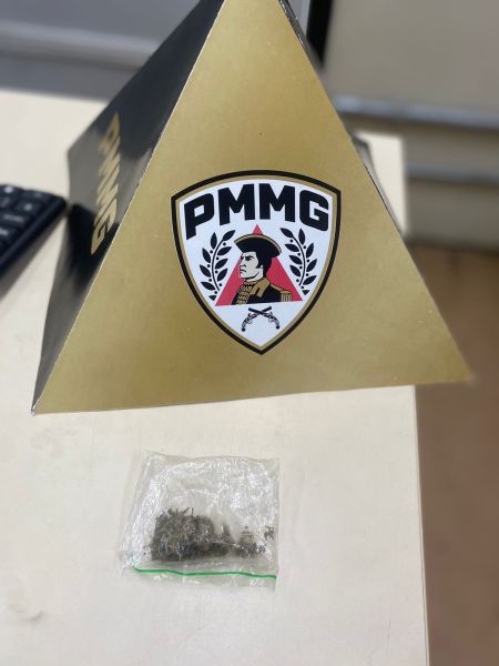 Uma caixa em formato de pirâmide com o logo da PMMG (Polícia Militar de Minas Gerais) e um saco plástico contendo uma substância vegetal em uma mesa.