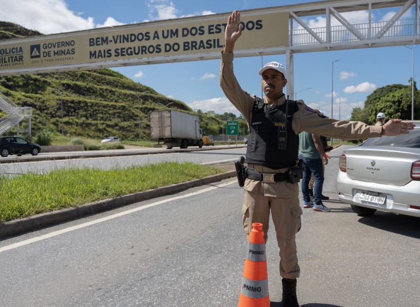 Um policial militar direcionando o tráfego em uma estrada, com um letreiro ao fundo que diz 'Bem-vindos a um dos estados mais seguros do Brasil'.